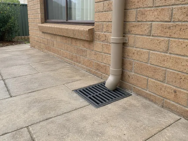 Stormwater grate beside a brick wall and downpipe on paved ground in Prairiewood
