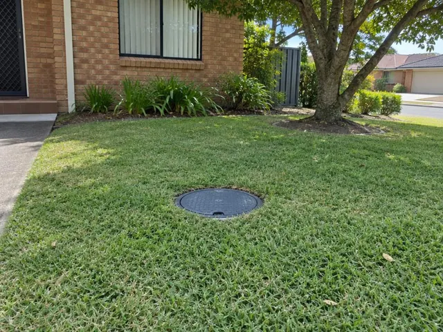 Lawn with a round capped sewer access cover beside a brick house in Bossley Park